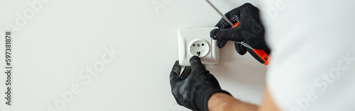 Close up of hands of male worker, professional electrician in protective gloves using screwdriver while installing new electrical socket outlet after renovation work