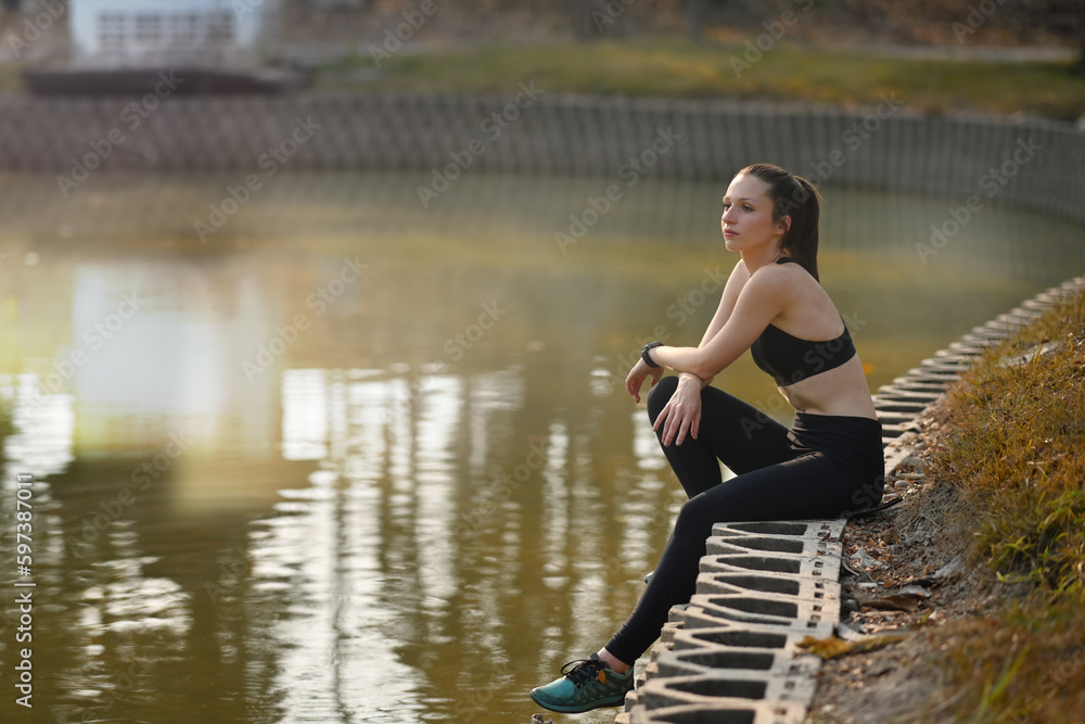 Fitness young caucasian woman sitting near river, resting after morning workout outdoor at summer park