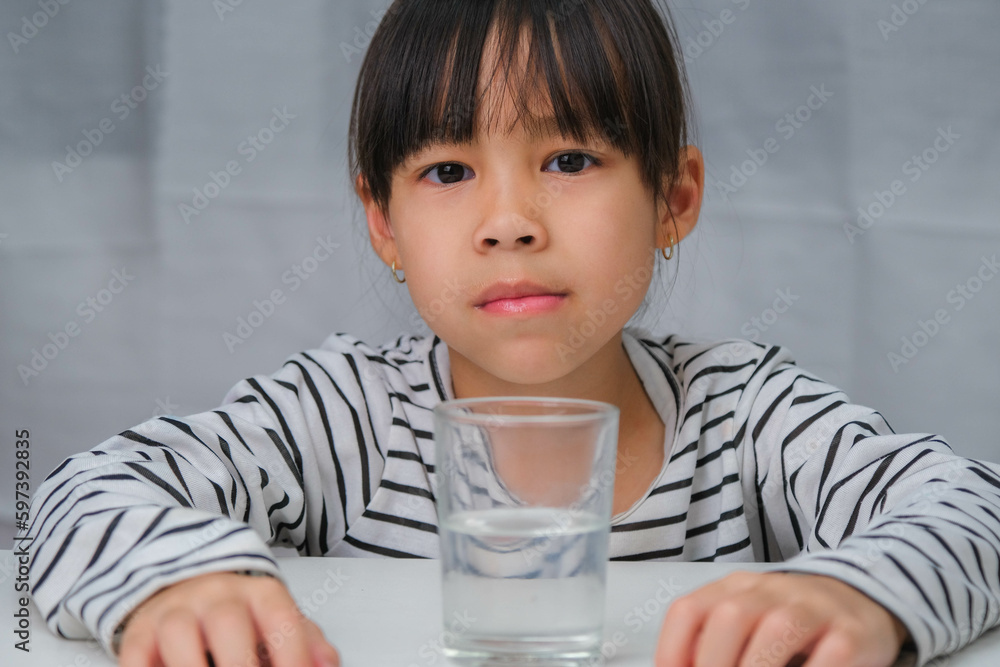 Cute little girl drinking water from glass on white curtain background. Healthy lifestyle and refreshment concept. Cute little girl drinking water from glass on white curtain background. Healthy lifestyle and refreshment concept.