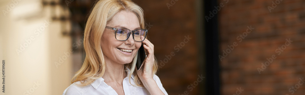 Morning. Cheerful middle aged woman with blonde hair and glasses holding a cup of coffee or tea while talking on her phone, standing at home