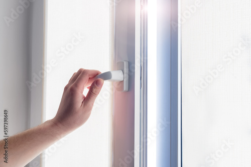 A man's hand holds a plastic window handle.Manual opening white plastic pvc window at home.A man opens a window, closes a plastic frame in his apartment. Roll-up blinds to protect against sunlight.
