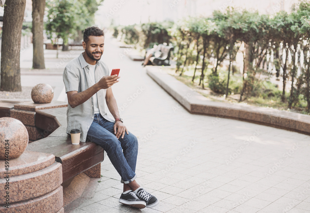 © kite_rin - Young student man using smartphone in a city. Smiling joyful guy summer portrait. Handsome man looking at mobile phone