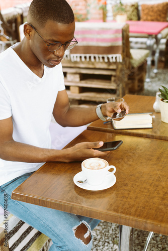 Afro american man with glasses sitting at table in cafe with cup of coffee on summer veranda, holding smartphone and entering number of credit card. Money, cashless payments, online shopping payment.