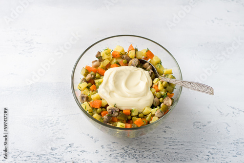 Glass bowl with vegetarian Olivier salad of potatoes, carrots, onions, pickles, mushrooms, green peas, dill with mayonnaise on a light blue background, top view. cooking stage