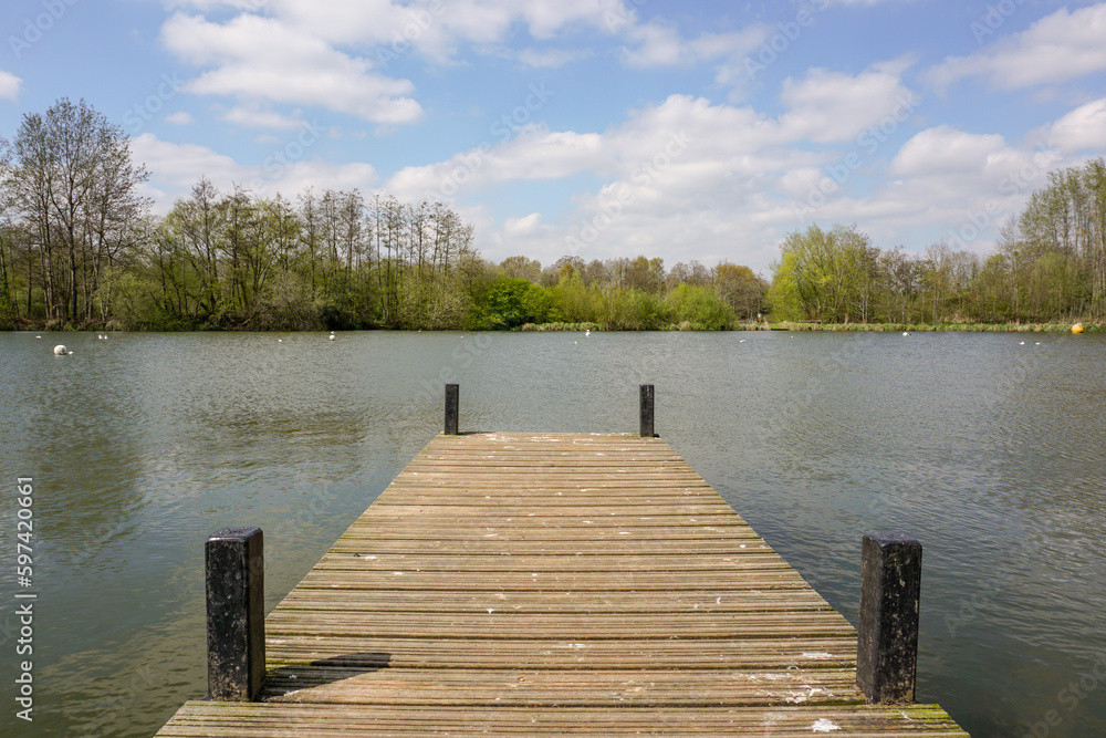 Naklejka premium view over wooden jetty over small lake with surrounding trees. peaceful scene in outdoor park space