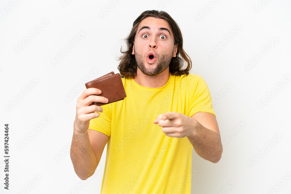 Young handsome man holding a wallet isolated on white background surprised and pointing front Young handsome man holding a wallet isolated on white background surprised and pointing front