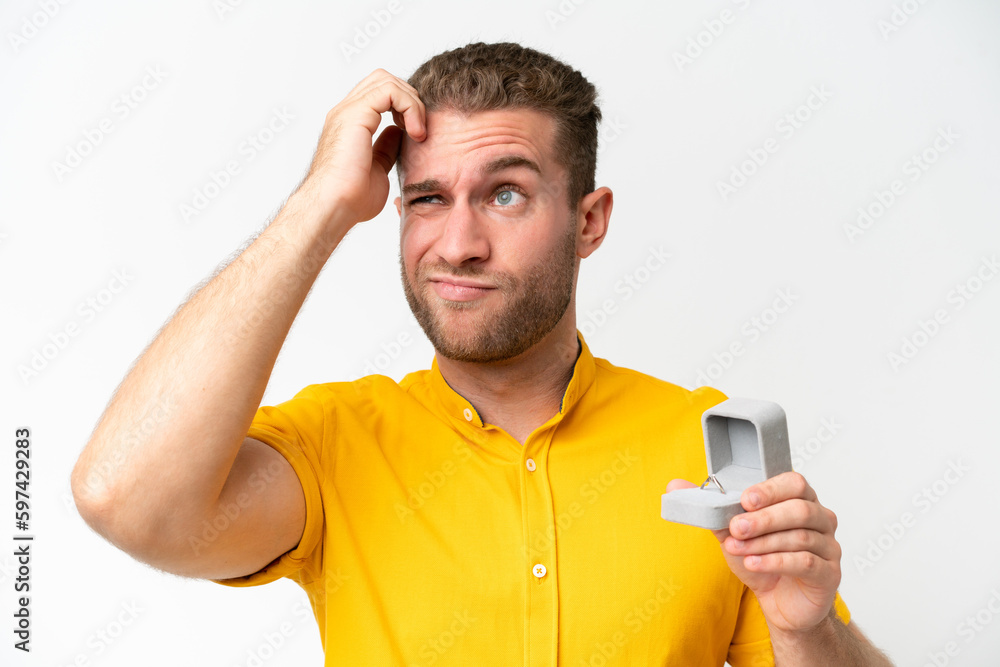 Young man holding a engagement ring isolated on white background having doubts and with confuse face expression Young man holding a engagement ring isolated on white background having doubts and with confuse face expression