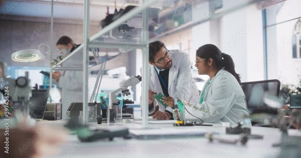 Two Diverse Multiethnic Colleagues Chatting in Laboratory, Working on a Circuit Board for a Computer. Young Indian Scholar Talking with a Scientist About Inserting Different Microchips and Conductors Two Diverse Multiethnic Colleagues Chatting in Laboratory, Working on a Circuit Board for a Computer. Young Indian Scholar Talking with a Scientist About Inserting Different Microchips and Conductors