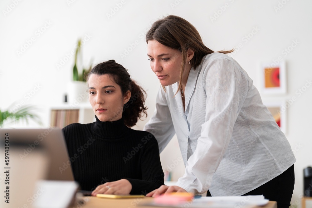 Young female coworkers in casual clothes sitting at table with laptop and looking at computer screen while working together on project in office Young female coworkers in casual clothes sitting at table with laptop and looking at computer screen while working together on project in office