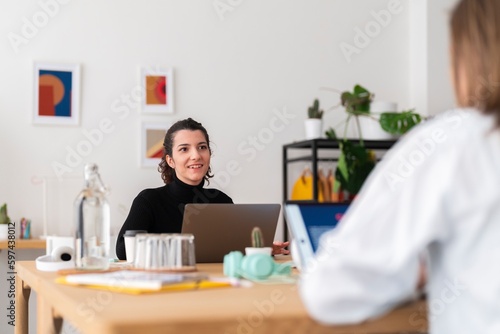 Two positive young female freelancers in casual clothes sitting at table with laptop and talking working on project photo