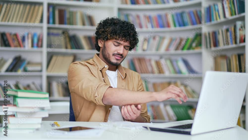 University student suffering from elbow pain while working on laptop while sitting in campus library space. The male massages the ligaments of the hand, rubs the painful joint, stretches the muscles