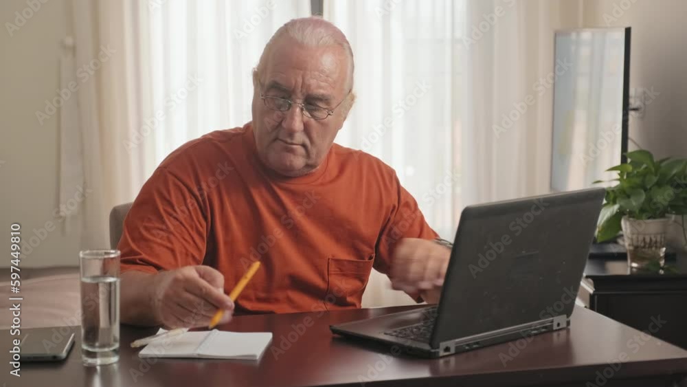 Medium shot of senior man in eyeglasses sitting at desk, working on laptop and making notes in copybook at home Medium shot of senior man in eyeglasses sitting at desk, working on laptop and making notes in copybook at home