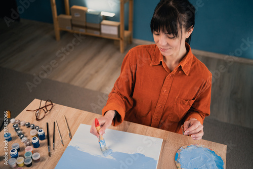 young beautiful caucasian woman sits at the table at home and paints a picture on canvas with acrylic paints. vacation and hobb concept. photo