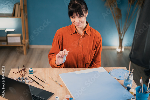a young beautiful caucasian woman sits at a table at home and paints a picture on canvas with acrylic paints, watches an online video lesson on drawing on a laptop. vacation and hobb concept photo
