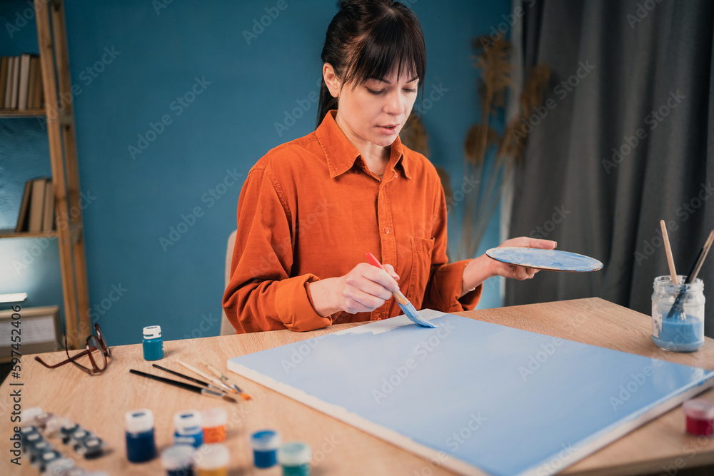 Beautiful woman in casual clothes paints with acrylic paints on canvas at home at the table using a brush. Beautiful woman in casual clothes paints with acrylic paints on canvas at home at the table using a brush.