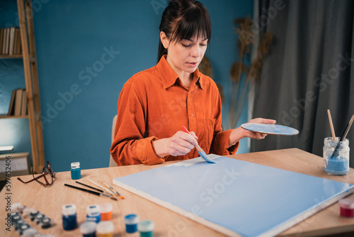 Beautiful woman in casual clothes paints with acrylic paints on canvas at home at the table using a brush.