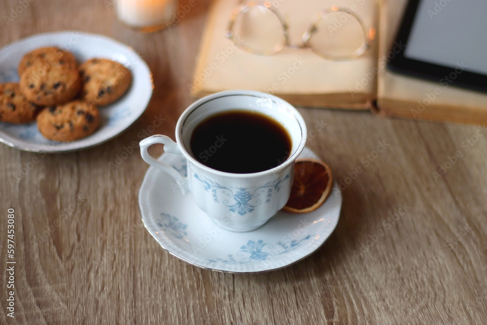 Cup of tea or coffee, plate of cookies, books, e-reader, pencil and lit candles on the table. Selective focus. Cup of tea or coffee, plate of cookies, books, e-reader, pencil and lit candles on the table. Selective focus.