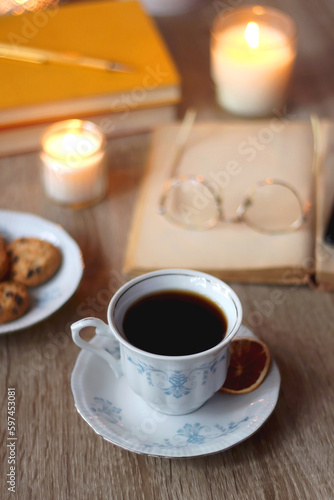 Cup of tea or coffee, plate of cookies, books, e-reader, pencil and lit candles on the table. Selective focus.