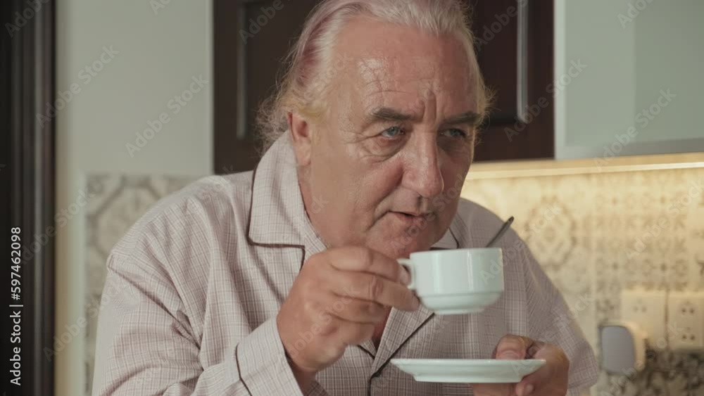 Medium close up shot of cheerful senior man in pajamas standing in kitchen, smiling and drinking freshly brewed coffee in morning at home Medium close up shot of cheerful senior man in pajamas standing in kitchen, smiling and drinking freshly brewed coffee in morning at home