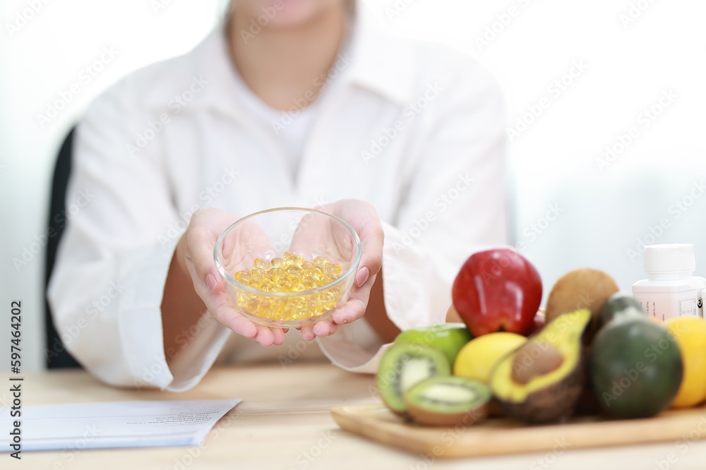 Woman doctor nutritionist hands in white shirt with omega 3, vitamin D capsules with green vegan food. The doctor prescribes a prescription for medicines and vitamins at clinic
