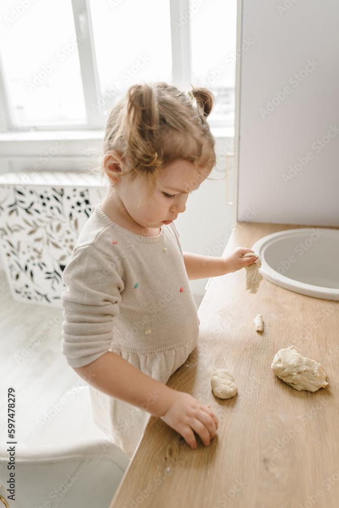 Fototapeta premium Little kid daughter having fun cooking pastries or pie on kitchen table top. Child knead dough, cooks pizza, bake cookies, dumplings, croissants for mother, father. Family preparation food on weekend.