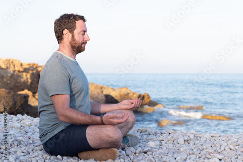 Profile view of a smiling man with eyed closed sitting in Sukhasana yoga pose on a pebble beach at sunset. photo