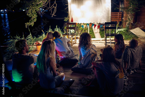 A group of people sitting on the floor in the bar on the river bank and enjoying night cinema. Night, summer, bar, river