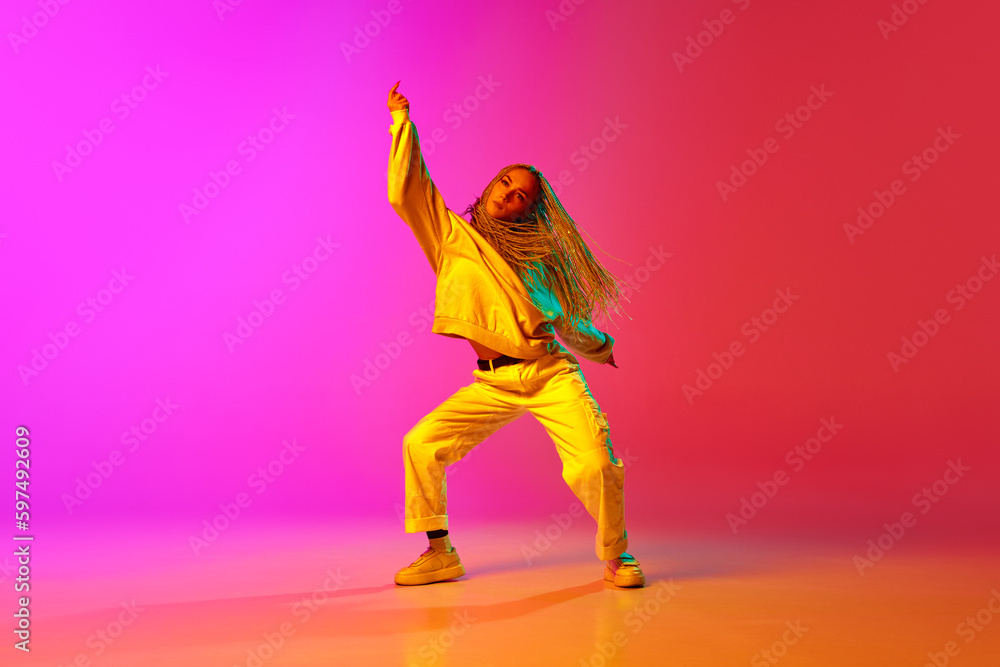 Portrait with one charming girl, dancer with dreadlocks wearing hip-hop style of clothes and dancing over gradient pink background in neon light