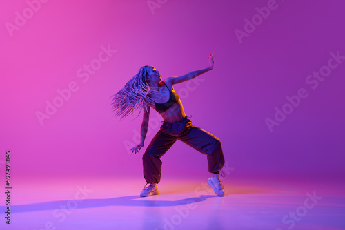 One young, attractive girl with dreadlocks dancing in street style over gradient purple neon background. Contemporary dance in motion