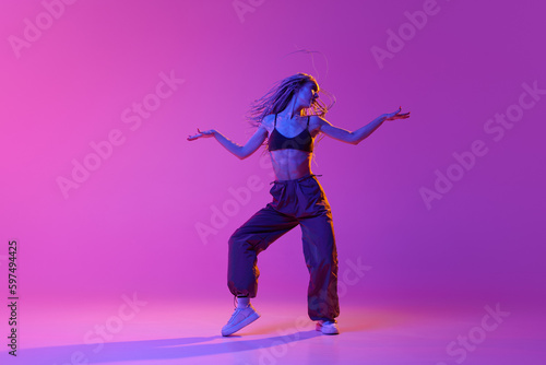 One young professional hip-hop dancer wearing stylish clothes dancing with inspiration over purple background in neon light. Beauty of movement