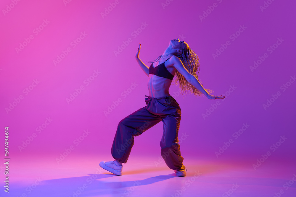 One attractive young woman, girl with pigtails dancing solo performance with pleasure over purple studio background in neon light. Freestyle