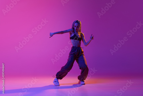 Portrait with modern young dancer wearing fashion clothes in motion over puple background in neon light. Liberty of dance
