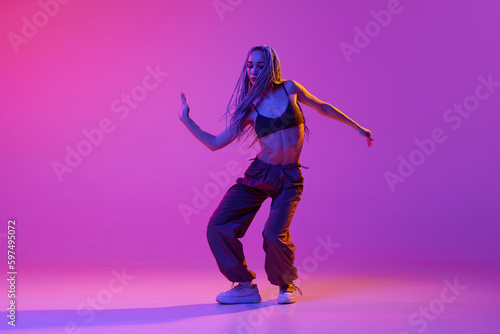 Young girl wearing street style clothes dancing with emotions over purple studio background in neon light. Hip-hop dance battle