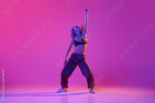 Young beautiful slim girl, modern dancer wearing hip-hop clothes dancing over gradient purple neon background. Contemporary dance style