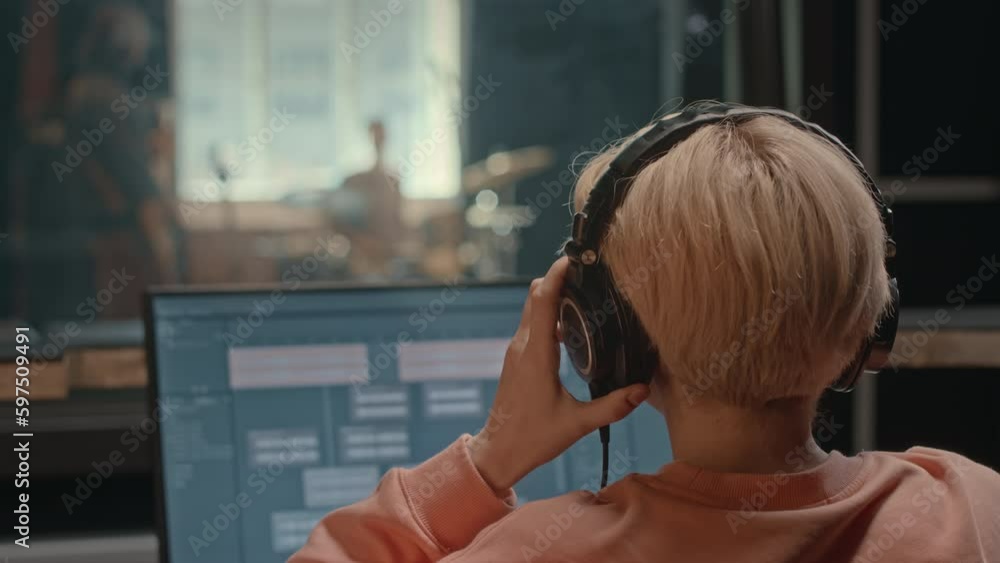 Back view of female producer in headphones sitting by workstation in sound studio recording music band playing in separate booth Back view of female producer in headphones sitting by workstation in sound studio recording music band playing in separate booth