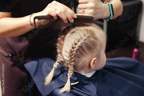 Barbershop. Hairdresser master woman make fashionable braids hairstyle for charming kid. Close up of hands barber makes hairdo for cute little blond girl child in modern hair salon. Copy text space