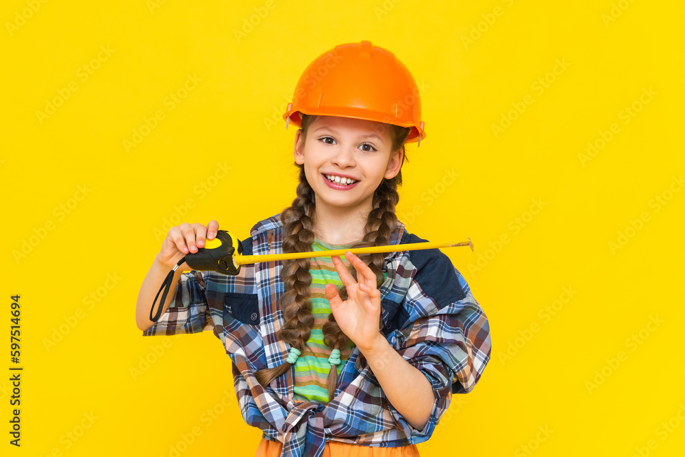 A happy young girl in a construction helmet holds a roulette tool. The child is preparing for repairs in the nursery. Yellow isolated background. A happy young girl in a construction helmet holds a roulette tool. The child is preparing for repairs in the nursery. Yellow isolated background.