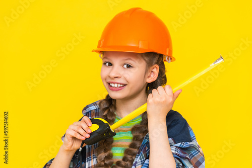 A happy young girl in a construction helmet holds a roulette tool. The child is preparing for repairs in the nursery. Yellow isolated background.