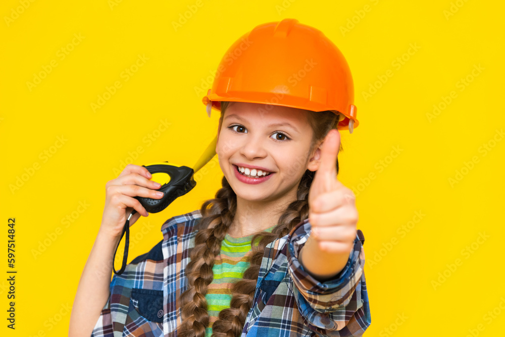 A teenage girl in a construction helmet shows a thumbs up sign and holds a tape measure. The child is an engineer. The concept of renovation in the children's room. A teenage girl in a construction helmet shows a thumbs up sign and holds a tape measure. The child is an engineer. The concept of renovation in the children's room.