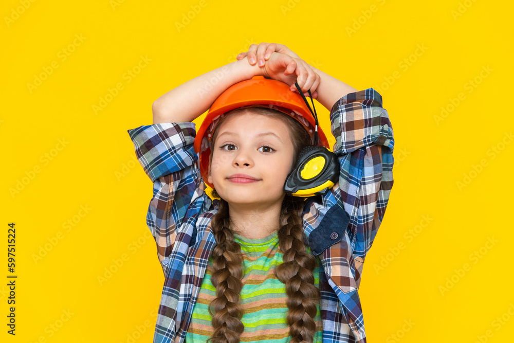 A child builder holds on to a construction helmet. A little girl is getting ready for repairs in the children's room and holding a tape measure. Yellow isolated background. A child builder holds on to a construction helmet. A little girl is getting ready for repairs in the children's room and holding a tape measure. Yellow isolated background.