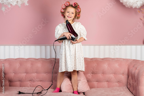 Little girl wearing hair rollers and white polka dot dress stands on the couch in the pastel pink living room, holds a hair dryer.