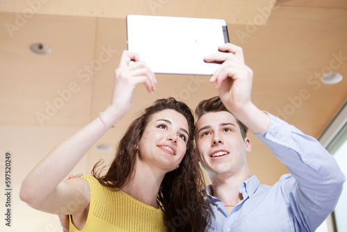 young and smiling couple taking a selfie with a white digital tablet. Boyfriend and girlfriend taking a photo using a tablet. Couple in love taking pictures together.