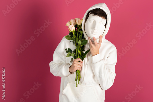 Suspicious young man in white hoodie, holds bouquet of flowers or roses and covering face with a mask. Secret admirer, Hidden identity concept. Isolated on pink background.