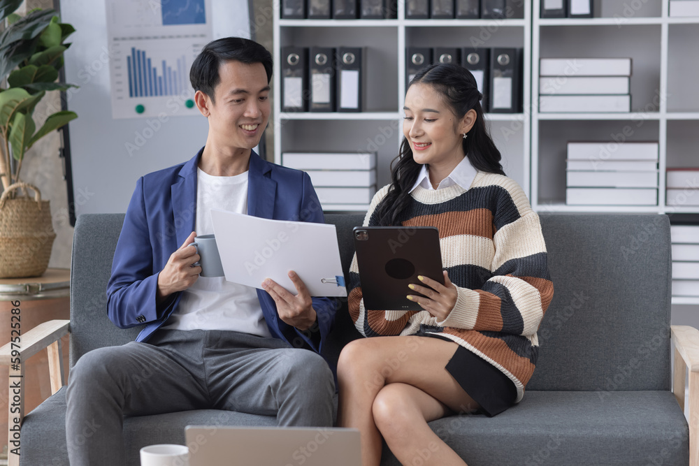 Young asian successful woman and man discussing paperwork and using a laptop together sitting on a sofa in a modern office. Young asian successful woman and man discussing paperwork and using a laptop together sitting on a sofa in a modern office.