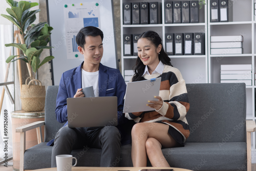 Young asian successful woman and man discussing paperwork and using a laptop together sitting on a sofa in a modern office. Young asian successful woman and man discussing paperwork and using a laptop together sitting on a sofa in a modern office.