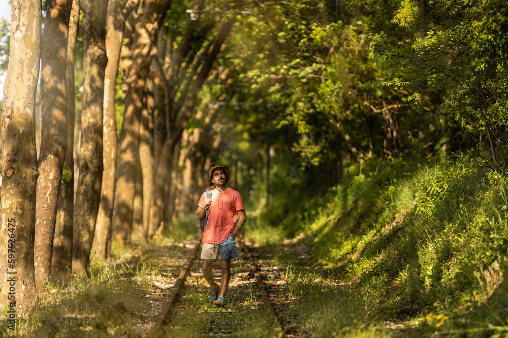 Backpacker man, walking along train tracks, observing Portugal's lush green forest in summertime. Wearing hat, pink t-shirt and short with sandals. Sunbeams.