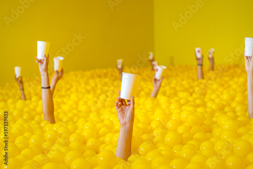 people's hands stick out of the pool with yellow balls, holding a white paper cup in their hand