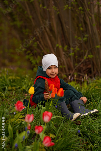 Cute child sitting in a meadow among tulips and holding a huge cone. Selective focus.