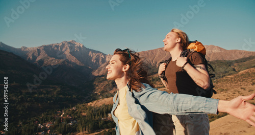 Man and woman hikers laughing with hands up, on a top of the mountain at sunset in mountains. Couple raising up hands on high rock in evening nature. Tourism, traveling and healthy lifestyle concept.