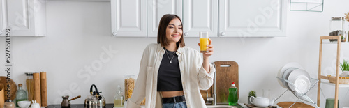 Positive brunette woman holding orange juice in kitchen, banner.